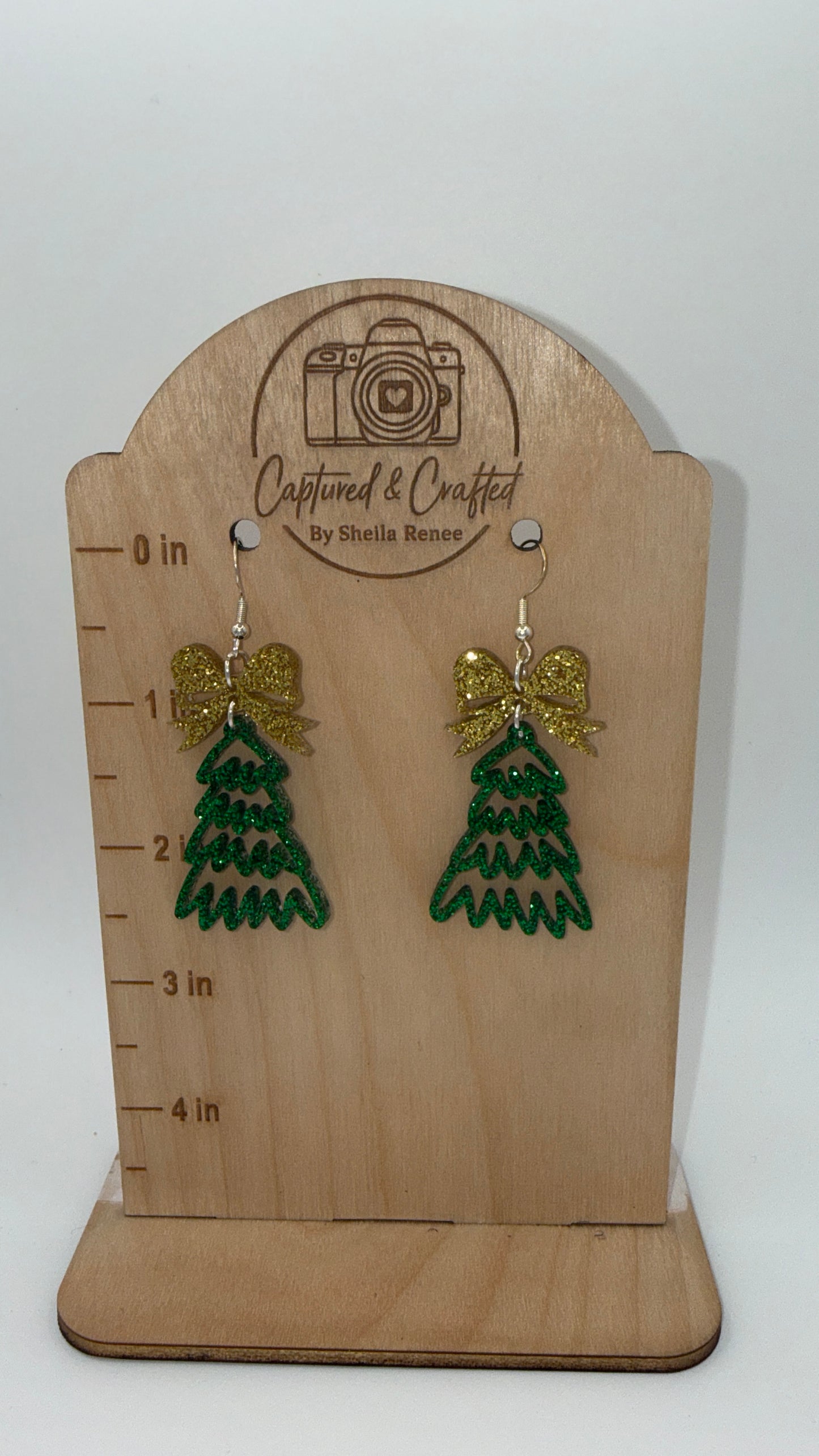 Green tree-shaped earrings with gold bows on a wooden display card with measurement scale.