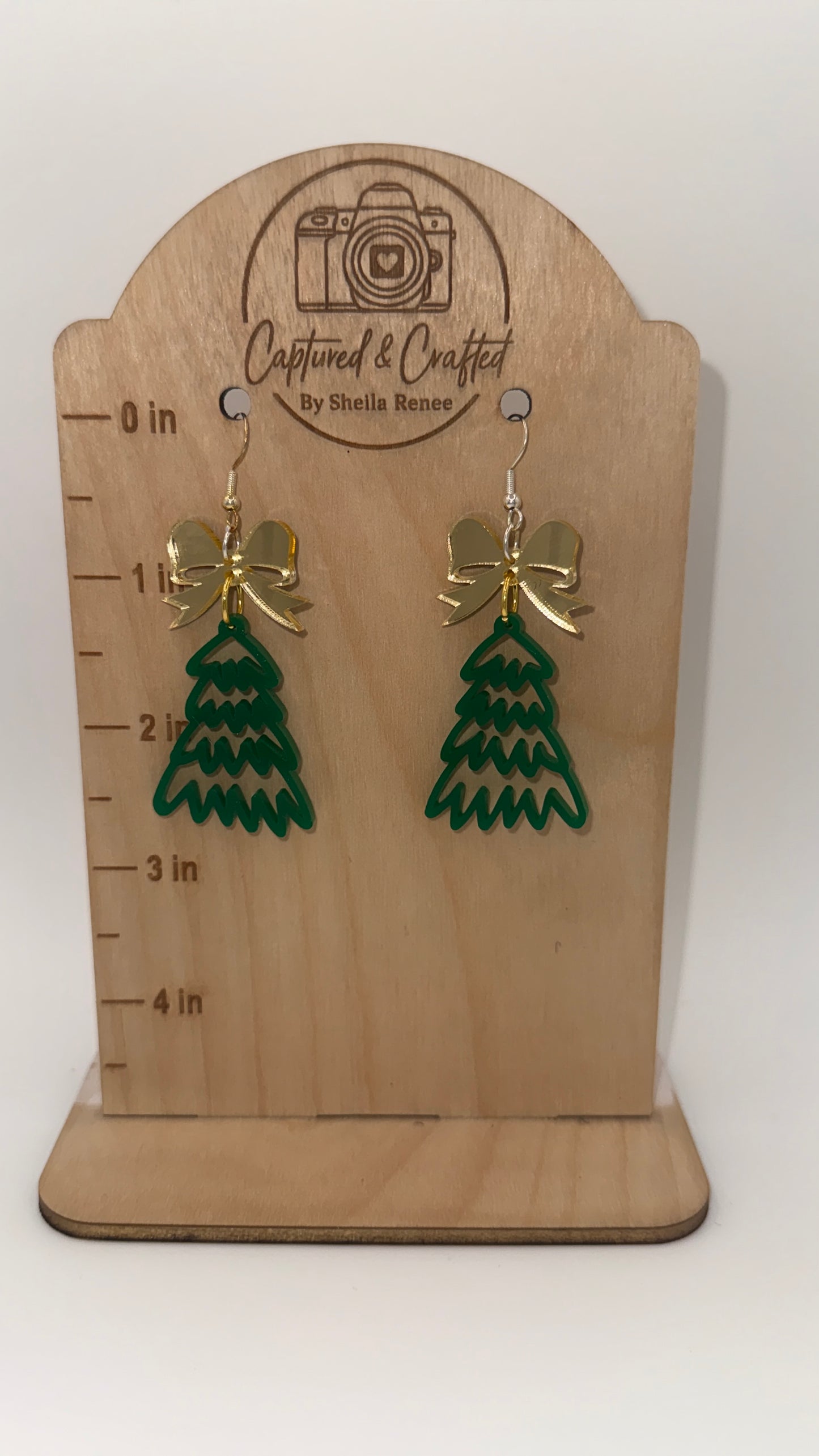 Green tree-shaped earrings with gold bows on a wooden display card with measurement scale.