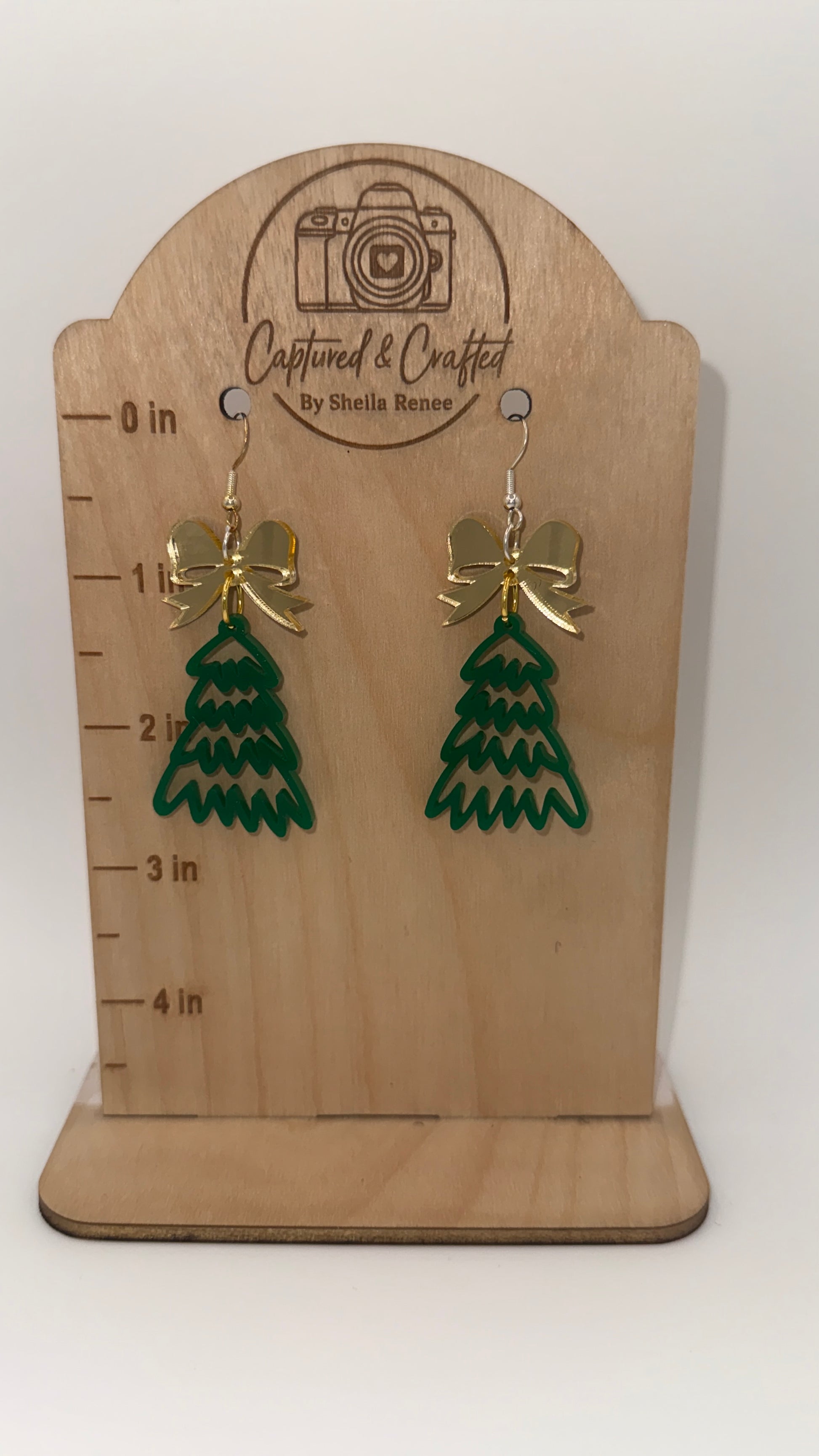 Green tree-shaped earrings with gold bows on a wooden display card with measurement scale.