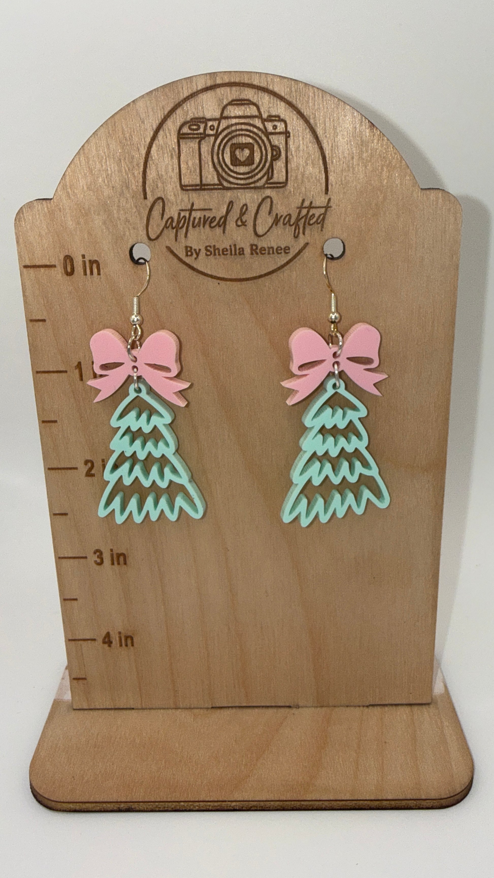Pink bow and mint green acrylic Christmas tree dangle earrings on a wooden display card.