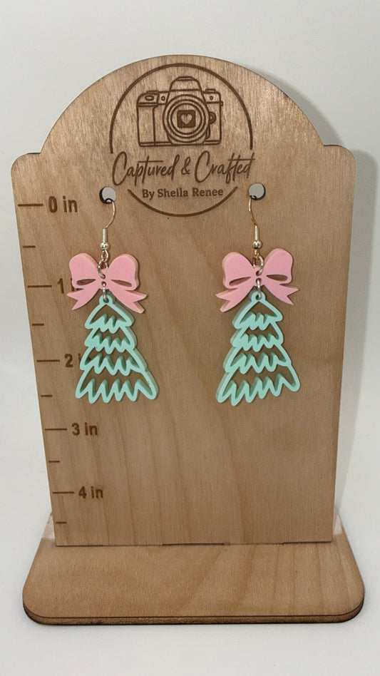 Pink bow and mint green acrylic Christmas tree dangle earrings on a wooden display card.